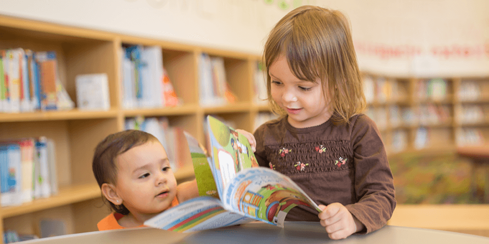 Photo of a baby and a toddler looking at a picture book in a library.