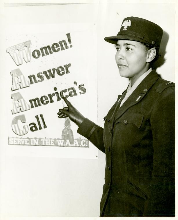 Captain in the WAAC,[African American Charity Adams, First Officer in the Women's Army Auxiliary Corps, standing in uniform and pointing to a poster that reads,