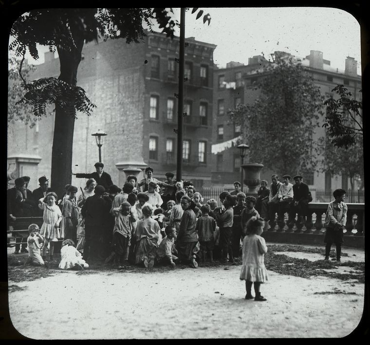 men listen along with children to story in the park, neighborhood buildings visible beyond, ca. 1910s., Digital ID 465199, New York Public Library