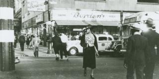 A busy 1939 Harlem street corner with an eatery are pictured. The camera is focused on an African American woman crossing the street, her head facing left. Two African American men in fedoras walk with their backs turned to the camera. 