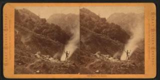 In a black and white stereoscopic photograph pair a figure is shown in the distance raising their arms before a tower of smoke on a grassy hillside, larger hills in the distance just barely visible through mist