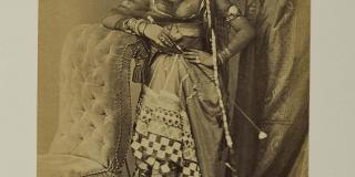 A sepia photograph of a young woman dressed in Indian garb. Her left hand rests on her hip and her right elbow rests atop a plush chair with a high back