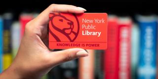 Hand holds a red NYPL library card with a lion logo and text that reads: New York Public Library, Knowledge Is Power. 