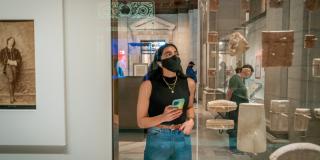 A woman with dark hair wearing jeans, a black sleeveless top, a black face mask, and earbuds holds her phone while looking at cuneiform tablets