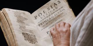 A light brown hand touches the pages of an open book with black type. The righthand page is titled Poesias Lyricas -- Primero Sueño.