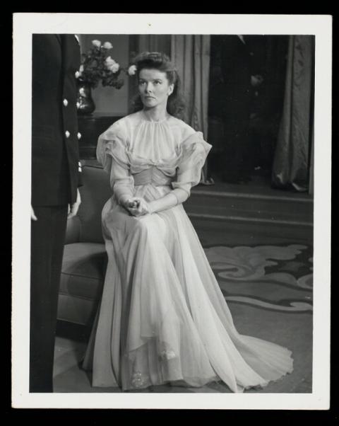 In a black and white photograph, Katherine Hepburn sits on the arm of a lounge chair