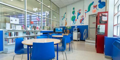 Interior of the High Bridge Library Teen Center, featuring work tables, books, and computers, with a colorful wall mural in the background.