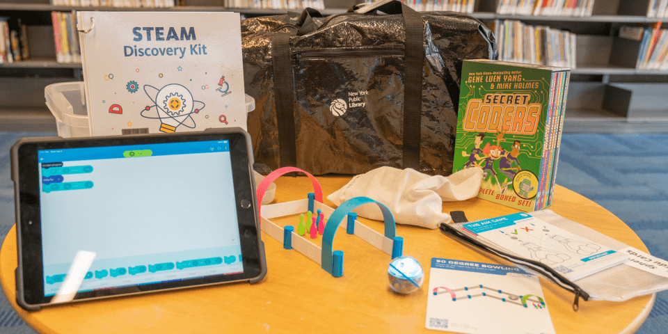 Photo of an NYPL Sphero Robotics Kit spread out on a table with items including: Sphero Mini robot and activity kit, iPad, and a six-book graphic novel series
