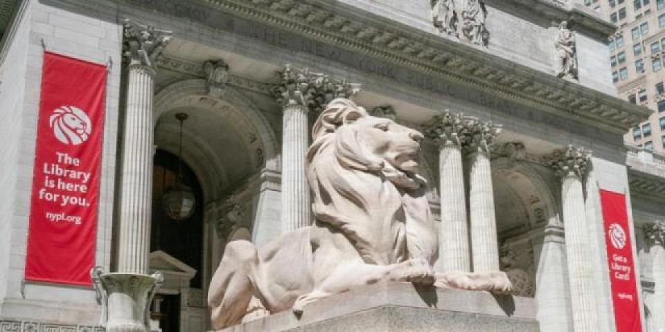 Marble lion statue outside of the Stephen A. Schwarzman Building, which is displaying two red banners that read: The Library is here for you.