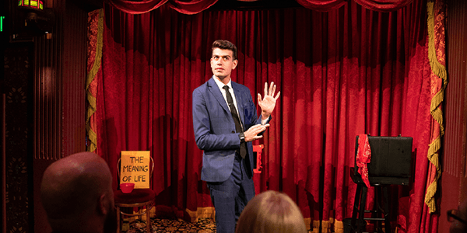 Man in suit on a stage with red curtains with arms raised, as if he was going to pull something out of his sleeve.