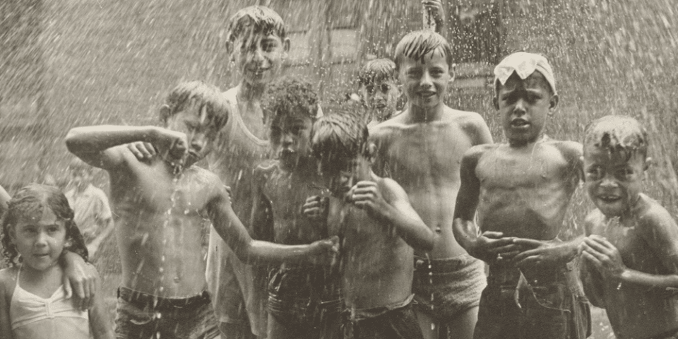 Historic black-and-white photo featuring a diverse group of children playing in the spray from a fire hydrant.