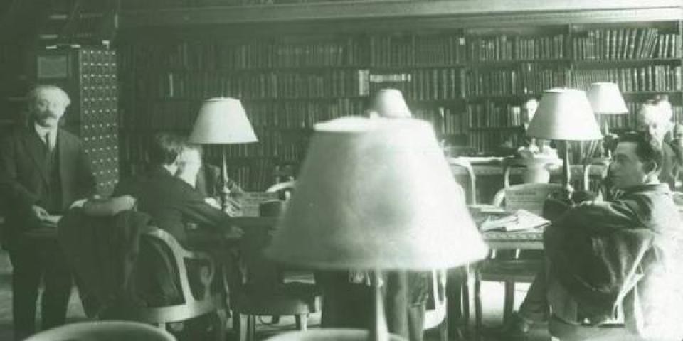 black and white photo of men sitting at tables in a library room lined with bookshelves