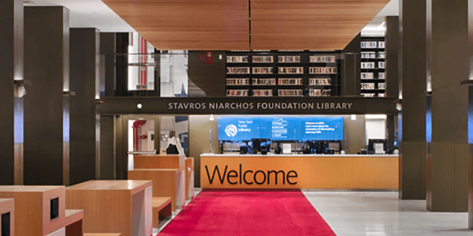 A red carpet unfurls into the Stavros Niarchos Foundation Library's lobby, decked out in rows of bookshelves with dark pillars and a wooden desk that reads Welcome.