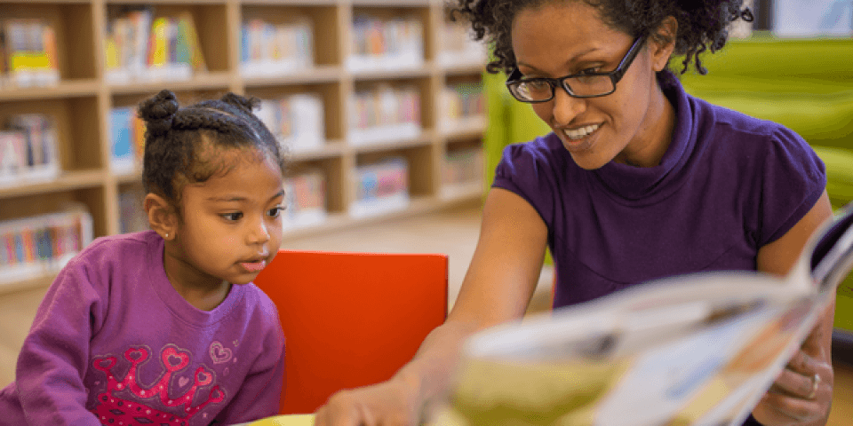Smiling adult reading a book to a toddler girl while sitting at a table in the Library.