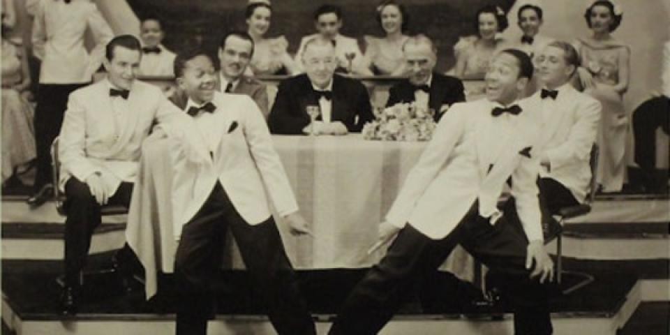 Two boys in white suits dance in front of a table of people.
