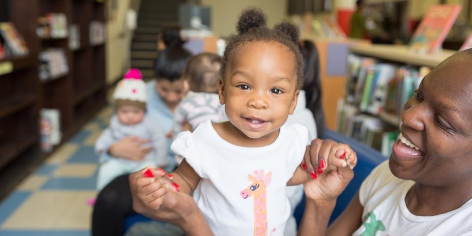 A one-year-old with buns in her hair smiles at the camera while being held by a caregiver in a library.