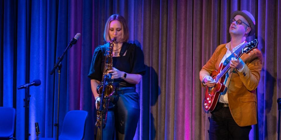 Performers Roxy Coss and Alex Wintz play sax and guitar at an event. 