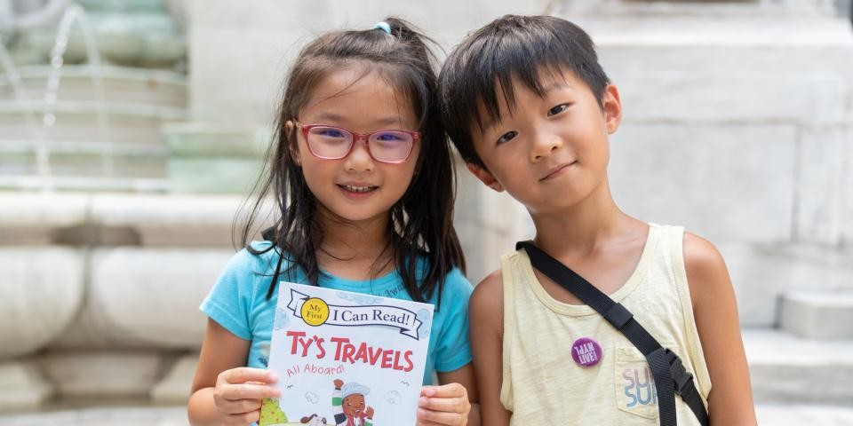 A young girl and boy smile and stand next to each other outside of the Stephen A. Schwarzman Building, holding a book. 