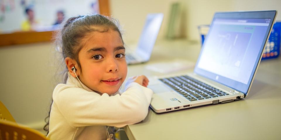 A young girl sits at a laptop and looks at the camera.