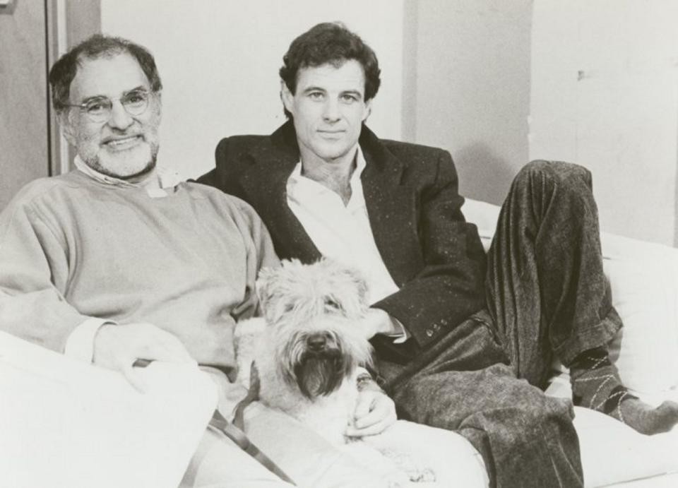Two men sitting on a couch, a younger man with a blazer, and an older man with a beard and a sweater. They have a dog between them