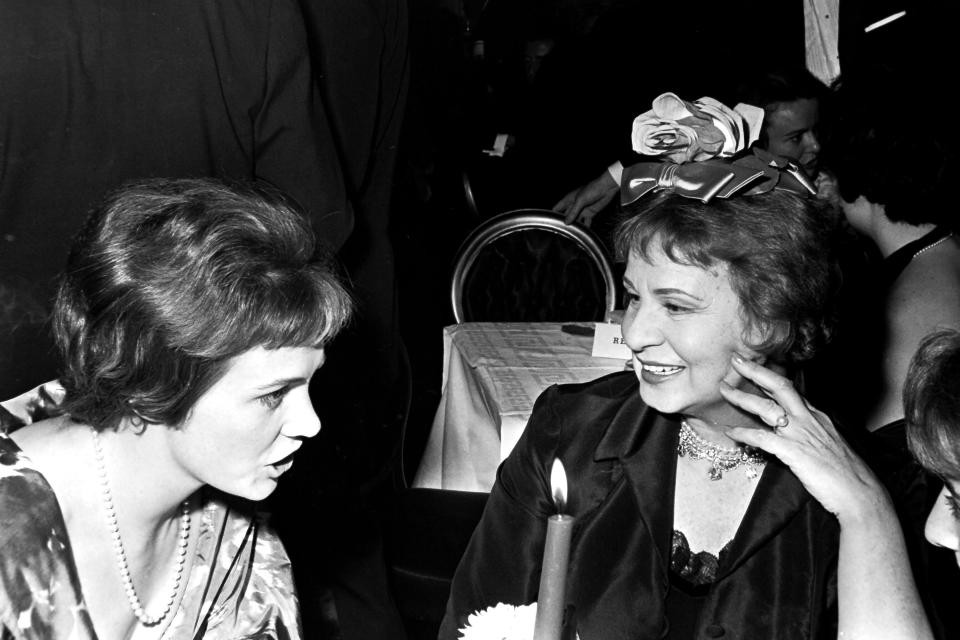 Two women speak at a dinner table. One is wearing a large rose and bow on the top of her head.