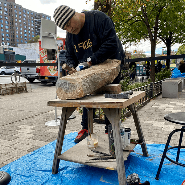 M. Scott Johnson is standing outside of the Schomburg Center holding a chisel and sculpting a gray colored stone.