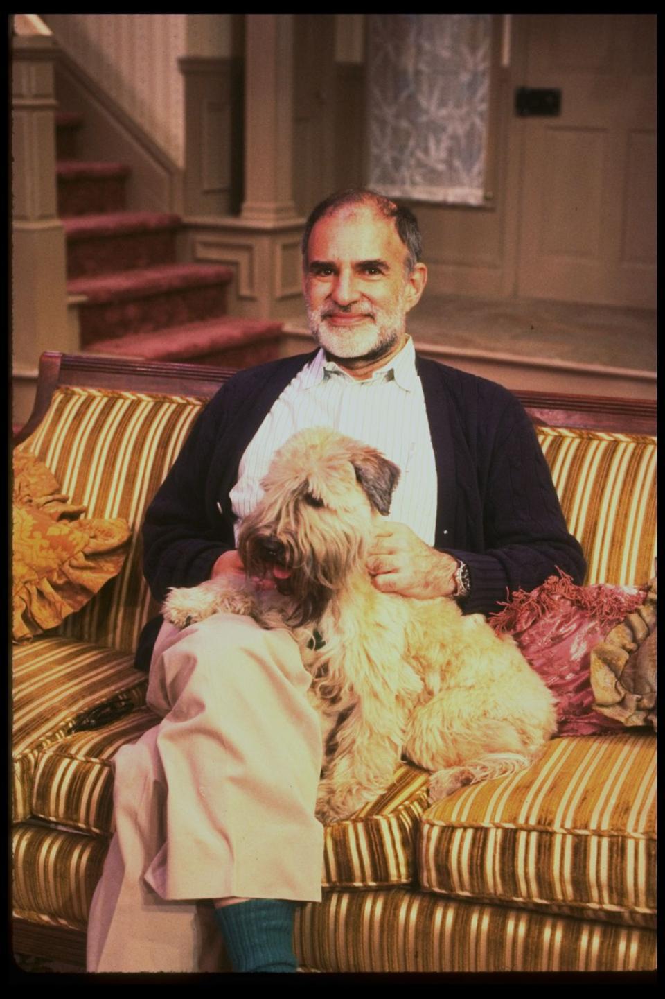 A bearded and bald man sits on a striped couch with a dog on his lap.