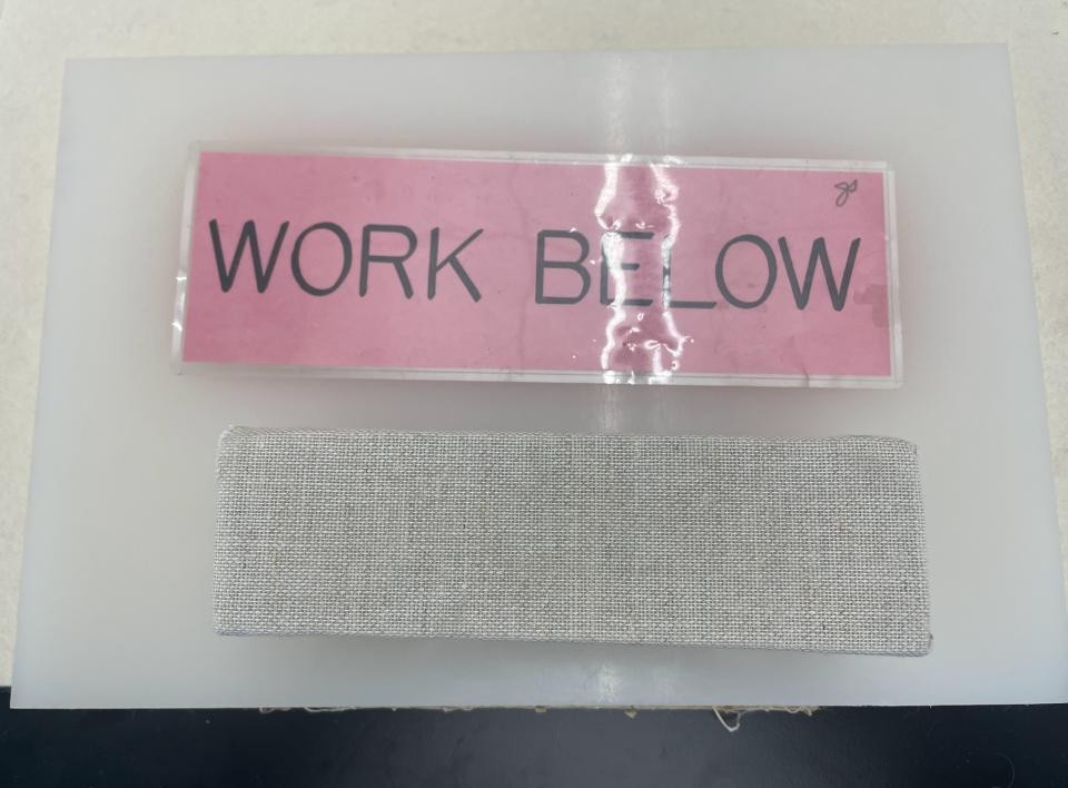 Laminated pink sign with the text Work Below. Under the sign is a brick-shaped, cloth-covered weight.