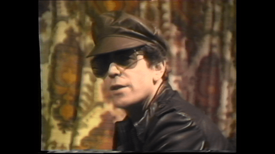 A video still of Lou Reed wearing leather jacket and hat, and a dark shades.