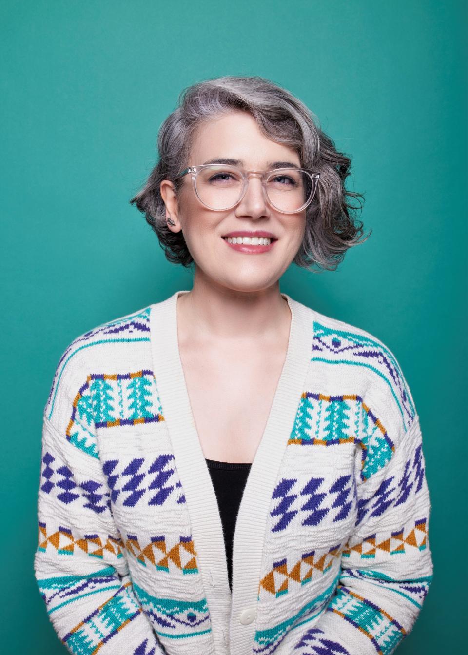 A young woman with gray hair and glasses and a cardigan sweater smiling before a teal wall