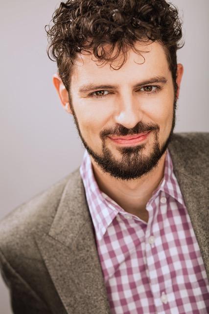 A man with a beard and short curly hair, wearing a plaid shirt under a gray blazer