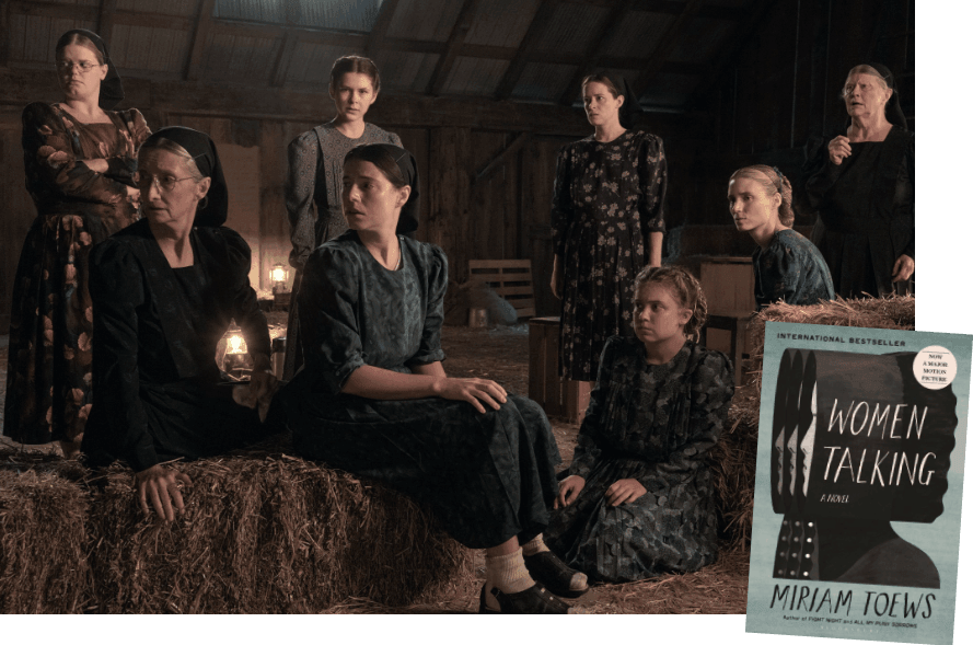 8 women in Amish dress sit and stand in a barn