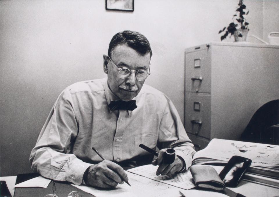 A man with a mustache, glasses, a pipe and pen in either hand, and a bowtie, writes at his desk
