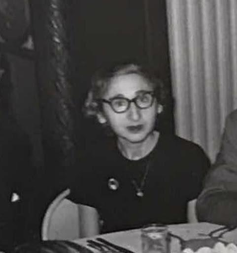 A black and white image of a woman wearing glasses sitting behind a table