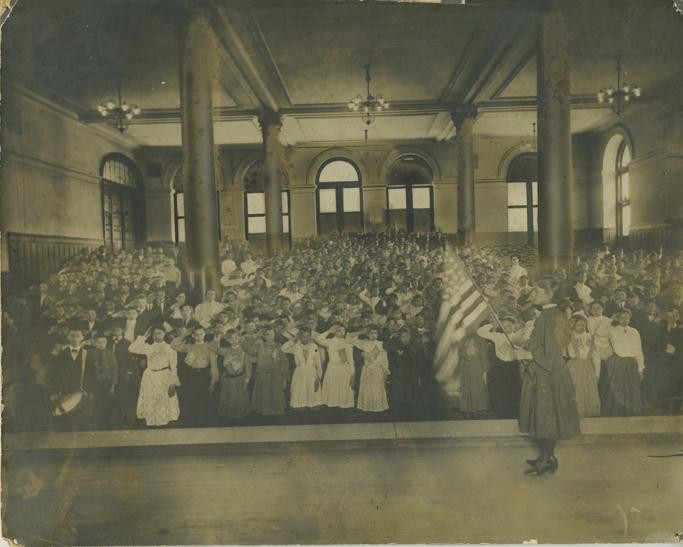 a crowded hall of girls and boys, all saluting, seen from a stage platform on which a girl holds a U.S. flag before them
