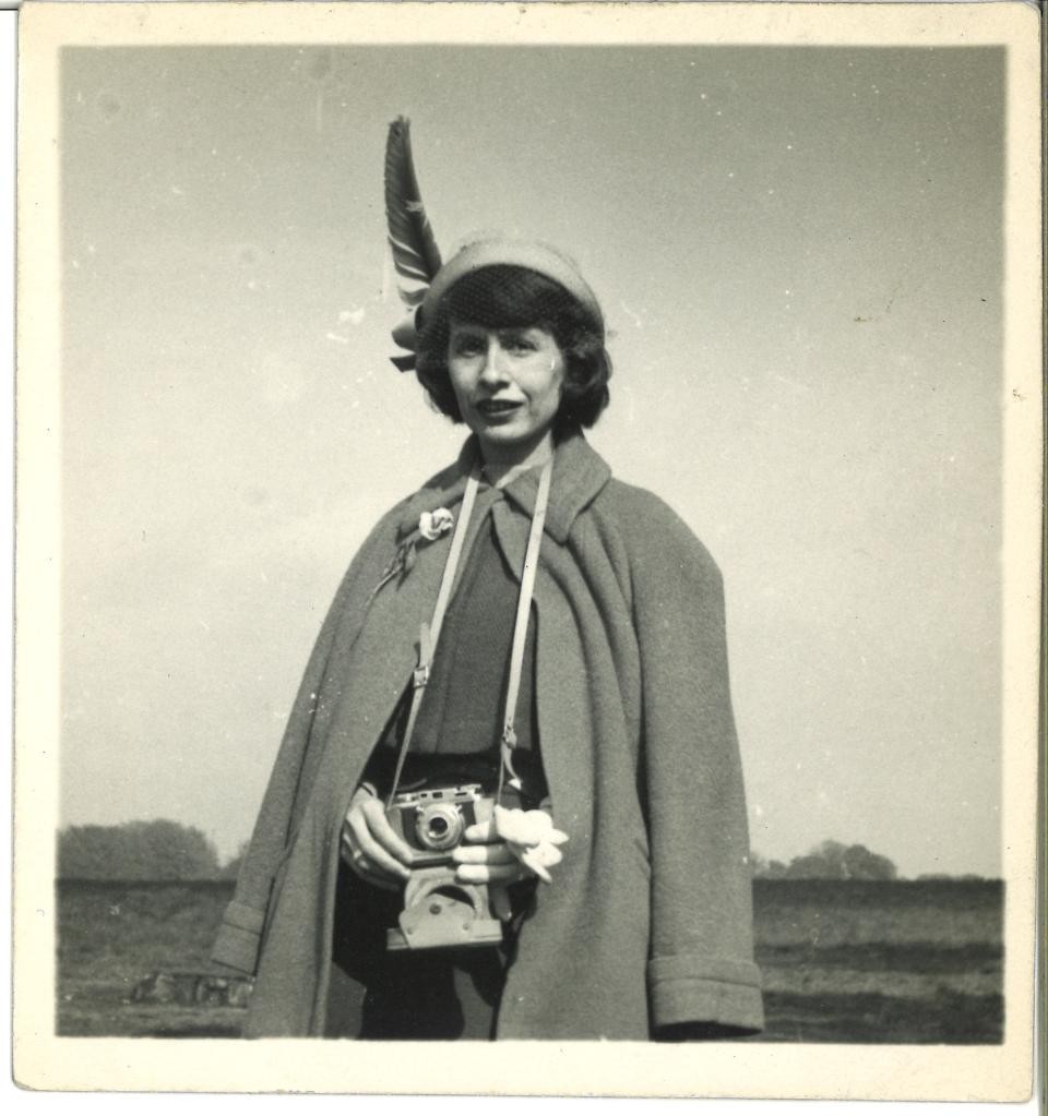 a black and white photograph of a woman wearing a coat and a hat with a large feather in it. She has a camera hanging around her neck.
