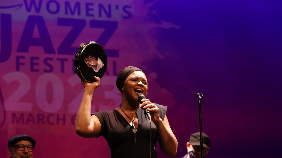 A woman is holding a microphone and signing. In the other hand, she has a tambourine. 