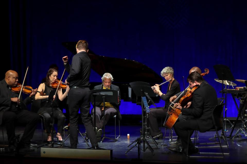 A group of musicians playing in a semi-circle. A conductor, back towards camera, is in the middle.