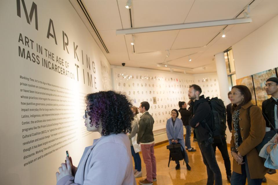 A group of people looking at a wall in the exhibition Marking Time. It has information about the exhibit listed. The wall is white. The letting is in black.