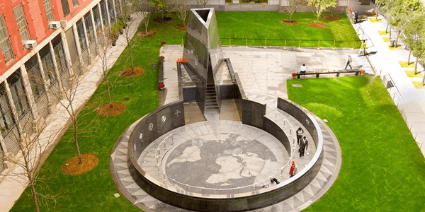 Aerial view of the African Burial Ground. There is a circular-shaped monument honoring the free and enslaved Africans and African-Americans whose remains are buried at the site. There is grass outside the structures, a cement pathway, and an office buildi
