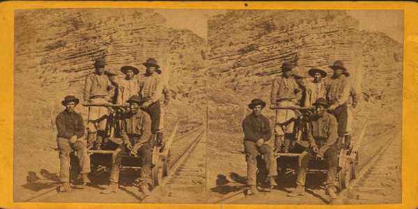 Yellowed photograph of workers of various races standing and sitting on a wagon along a line of railroad tracks.
