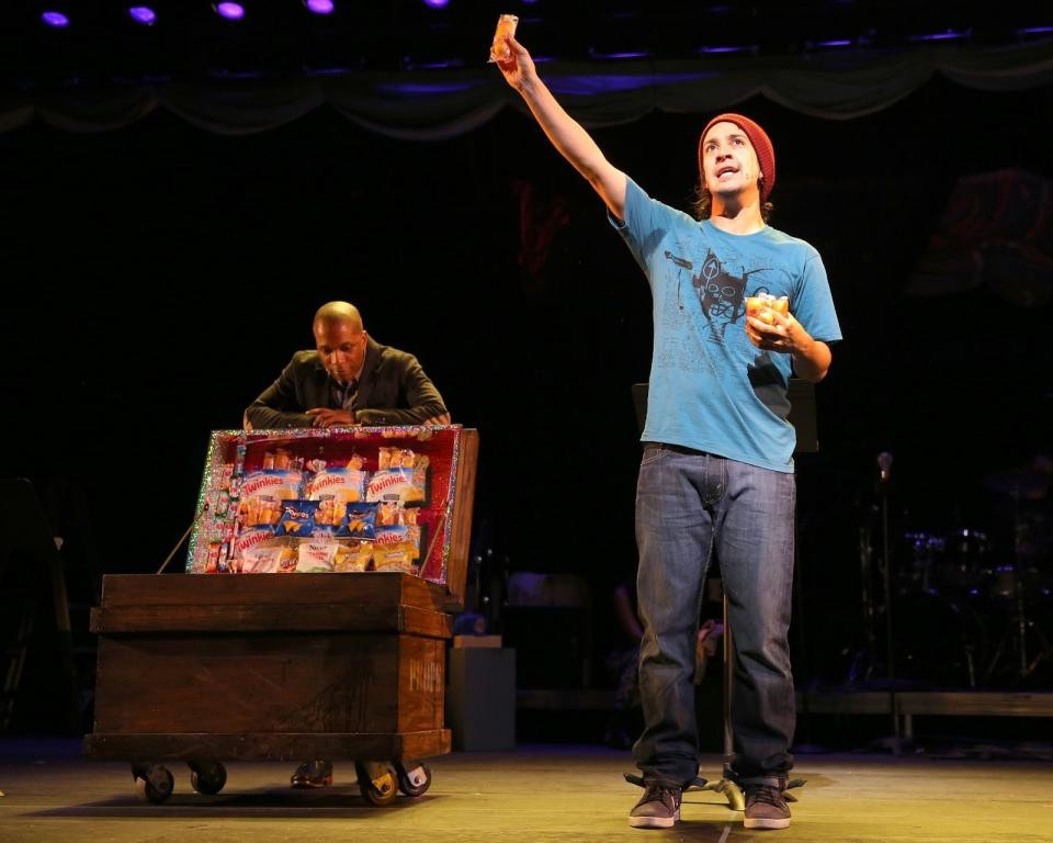 LIn Manuel Miranda raises a Twinkie in the air while Leslie Odom jr. looks at a cart of snacks in the background.