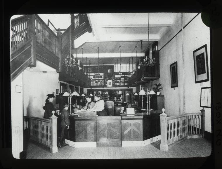 Black-and-white image of librarians at a large wooden reference desk assisting adults and children