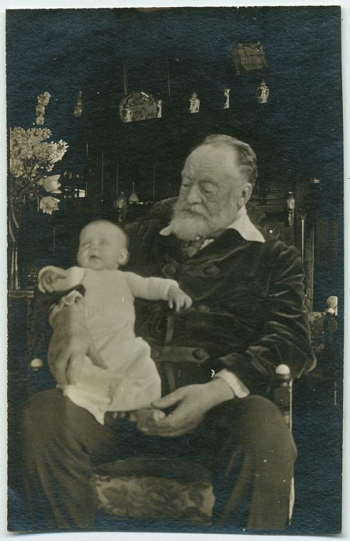 Historic portrait of older man sitting in chair holding infant. 