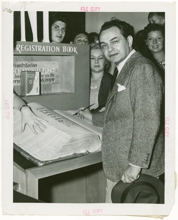 A rare historical image that displays a celebrated actor leaving his signature at the registration desk of the Jewish Palestine Pavilion. The picture also demonstrates a monumental size of the manuscript.