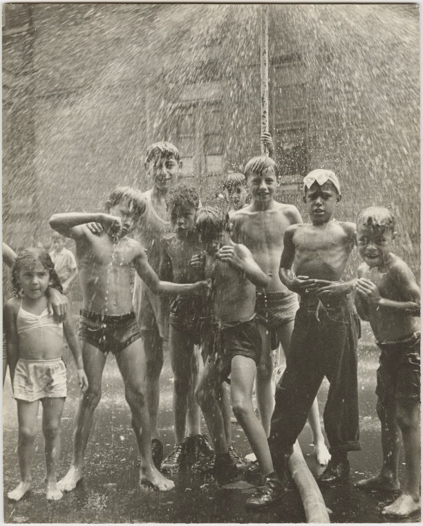 A group of shirtless children stand underneath a sprinkler in an East Harlem street; one of the children squares up with fists at the photographer while the others smile, looking directly at the photographer.