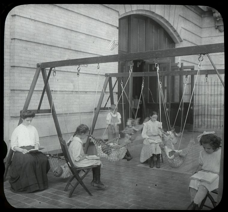 Women and girls reading near swingset where younger children are suspended in hammock like swings