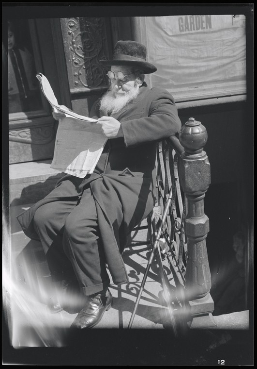 older man with a long white beard sitting on a stoop reading a Yiddish newspaper