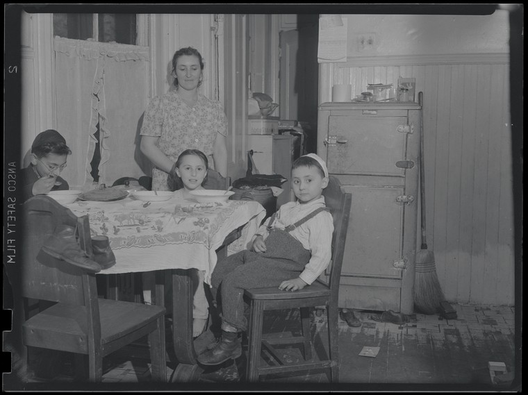 three children sit at a kitchen table while their mother stands nearby