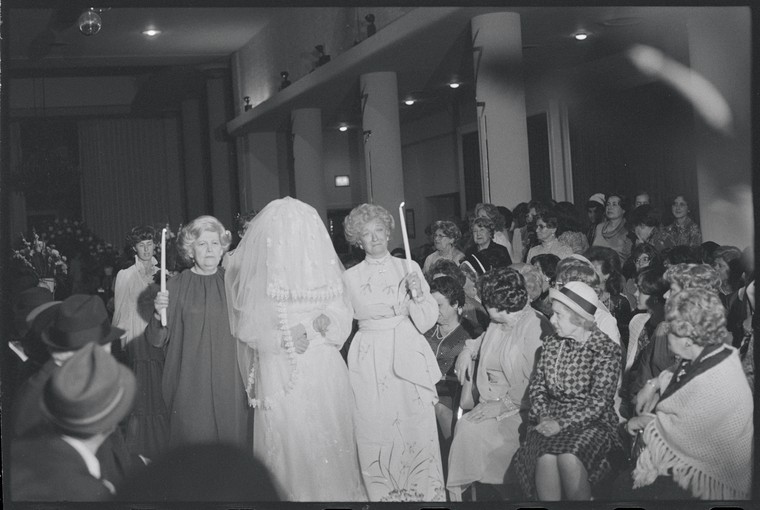 woman in white wedding dress and veil holding hands with two older women being led down an isle of a synogogue